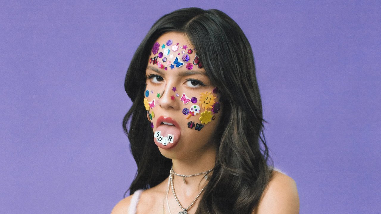 Olivia Rodrigo zieht die Zunge heraus, mit Gesichts- und Zungenaufklebern, die das Wort buchstabieren