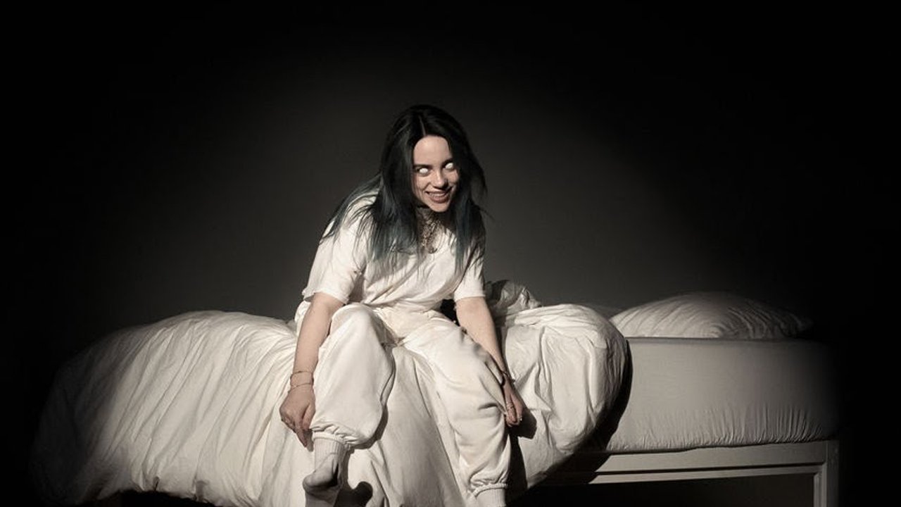 Billie Eilish sitzt auf einem weiß gekleideten Bett