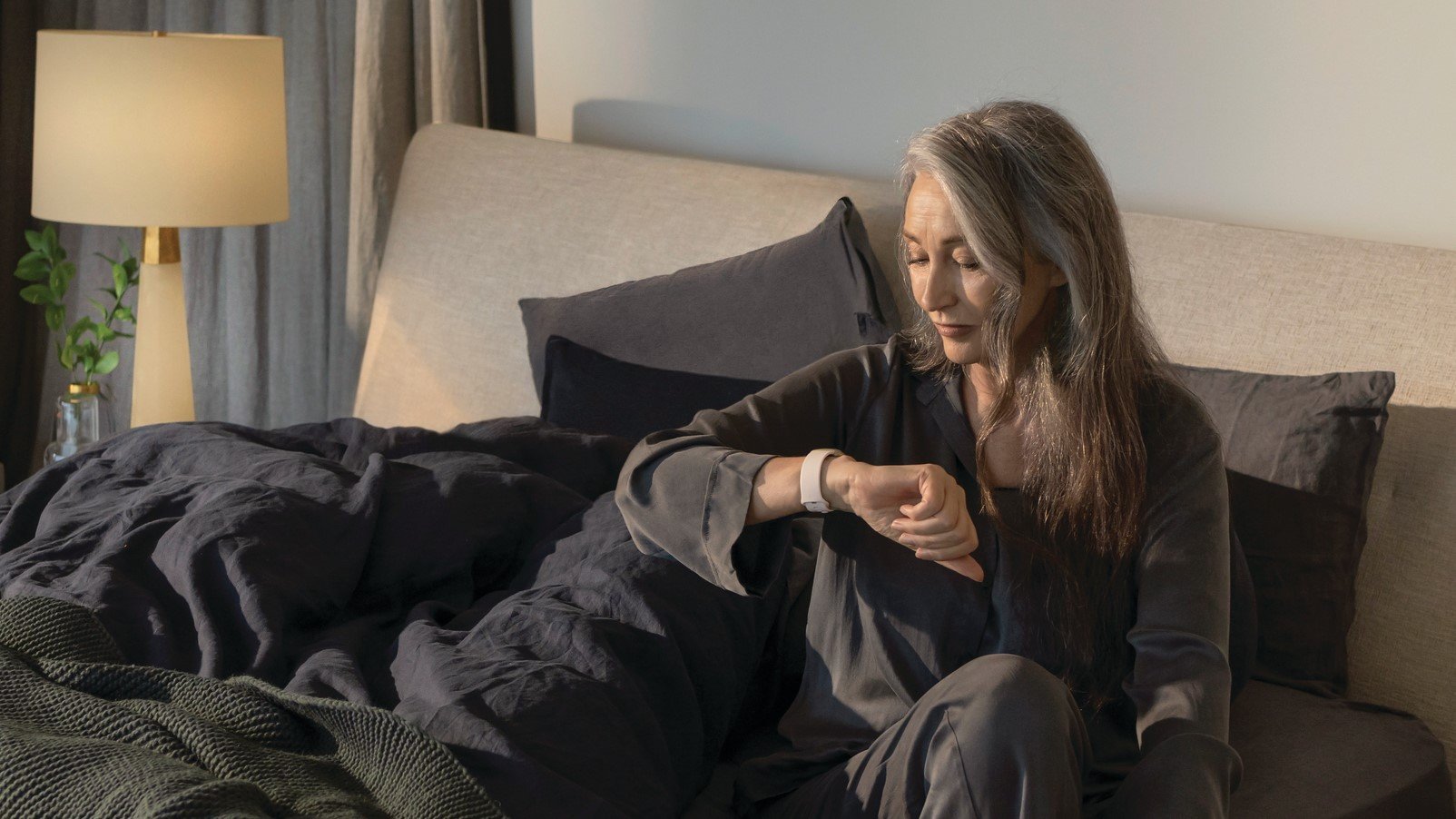 Женщина сидит на кровати и проверяет свои часы Fitbit Sense