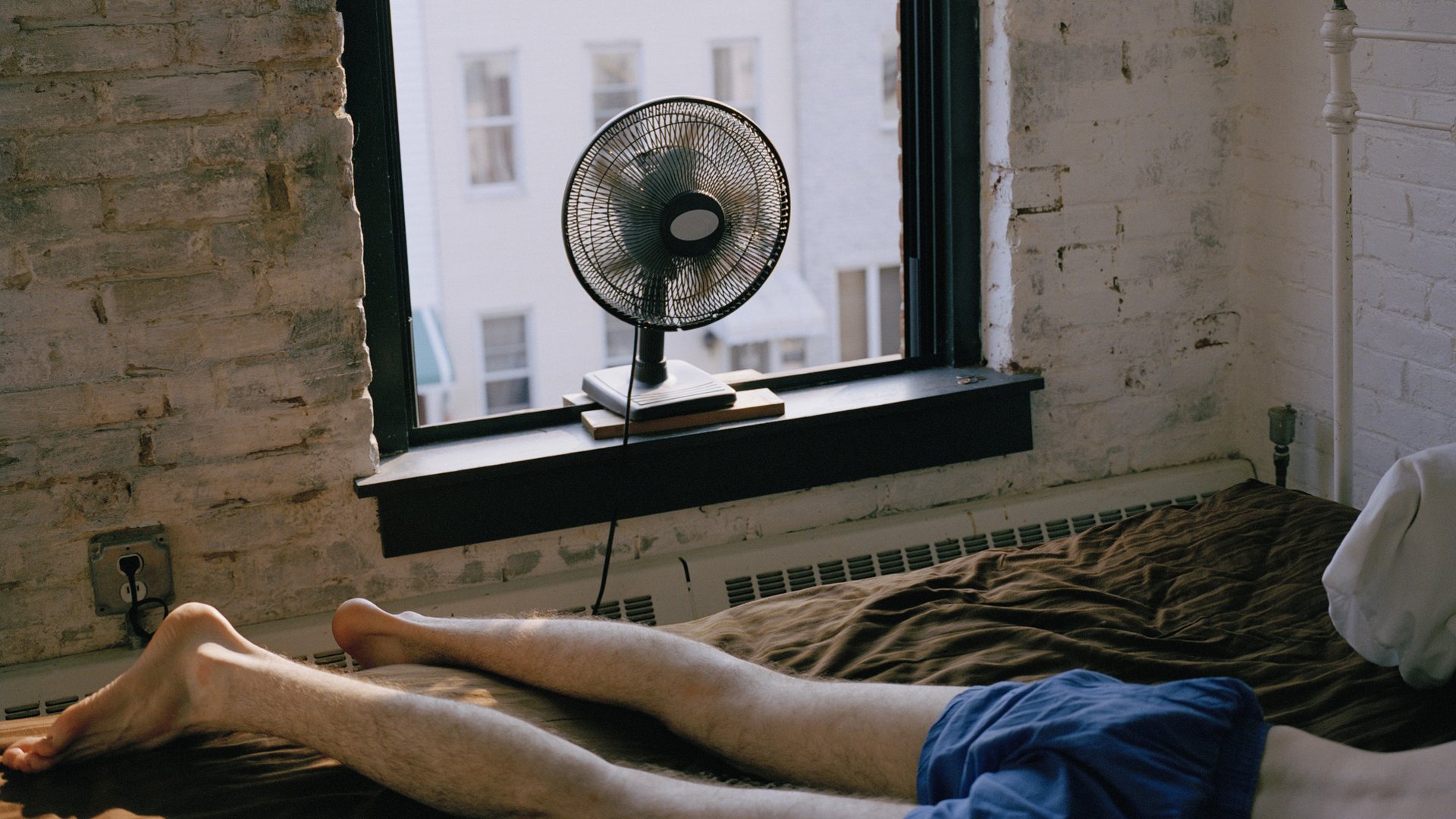Un uomo in pantaloncini blu dorme sul piumone con un ventilatore che soffia aria da una finestra aperta