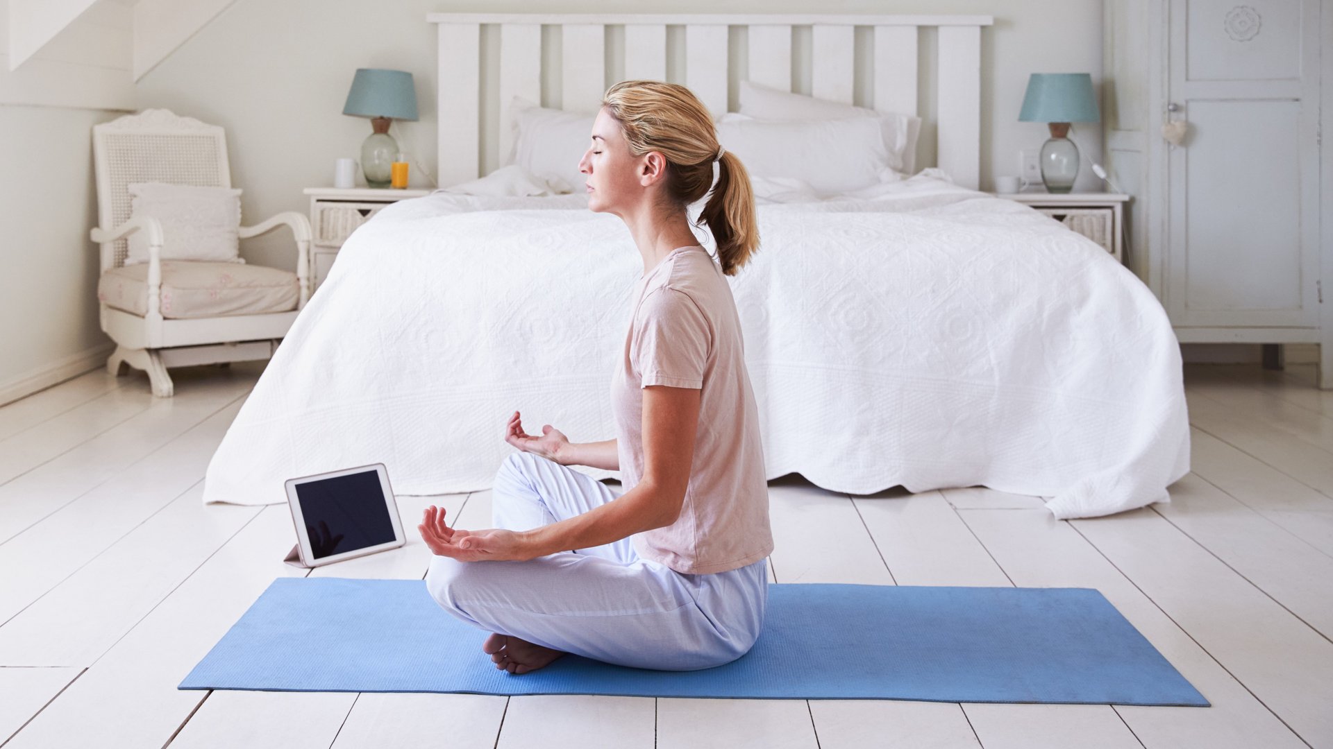 Une femme utilise une application de méditation assise sur un tapis de yoga bleu devant son lit blanc