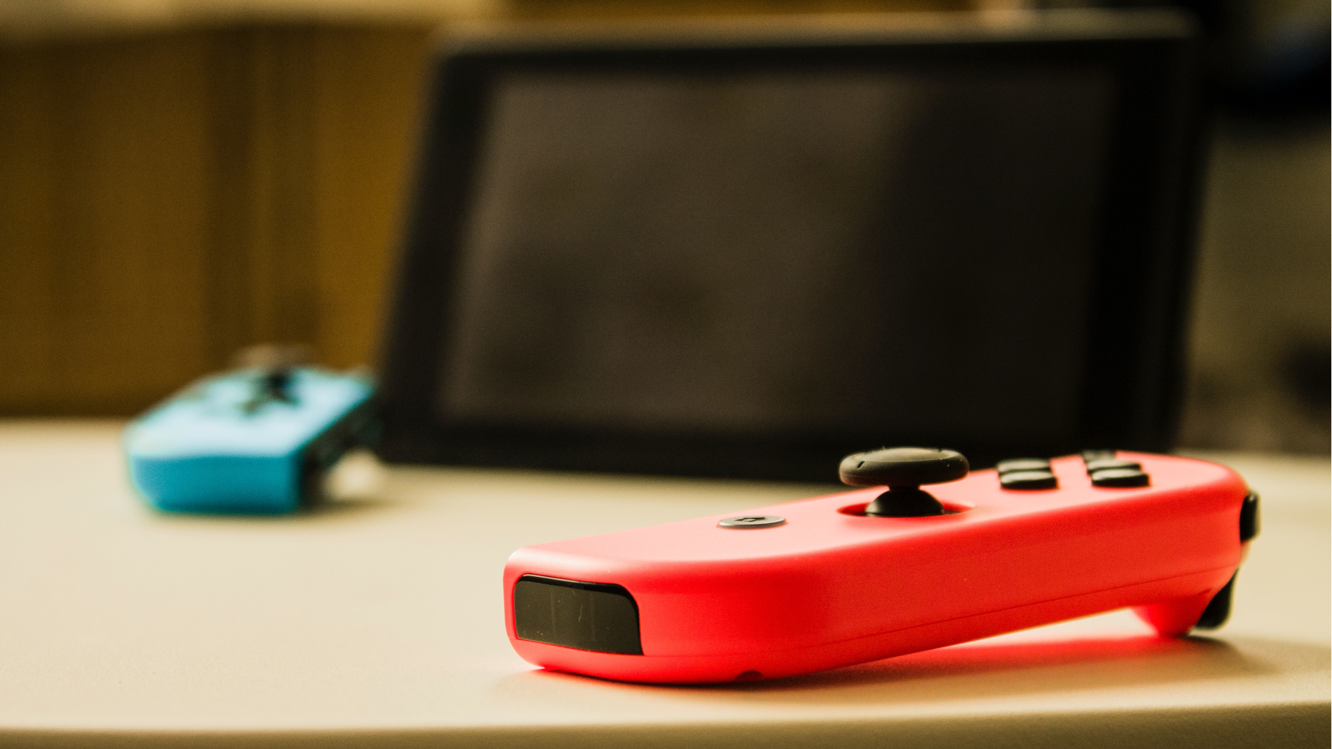 Un Joy-Con rojo neón sobre una mesa en primer plano, con una pantalla de Nintendo Switch y un Joy-Con azul descansando sobre la mesa en el fondo, ligeramente desenfocado