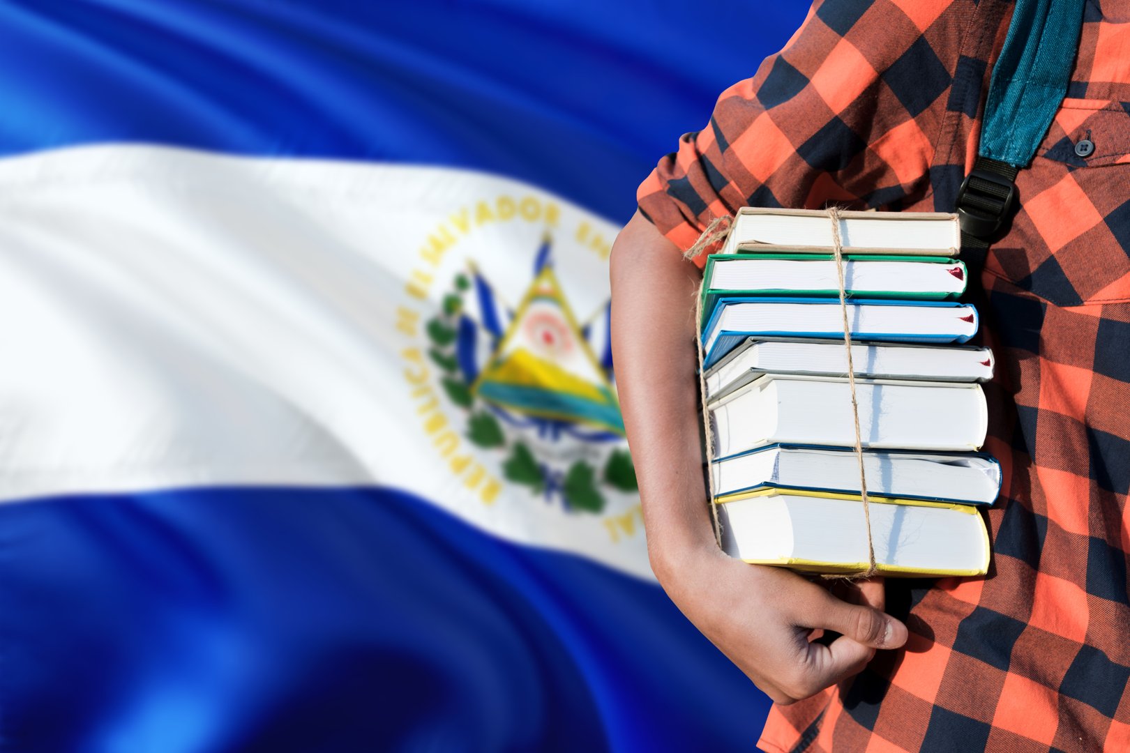 Un estudiante sostiene una pila de libros bajo el brazo contra el telón de fondo de la bandera salvadoreña.
